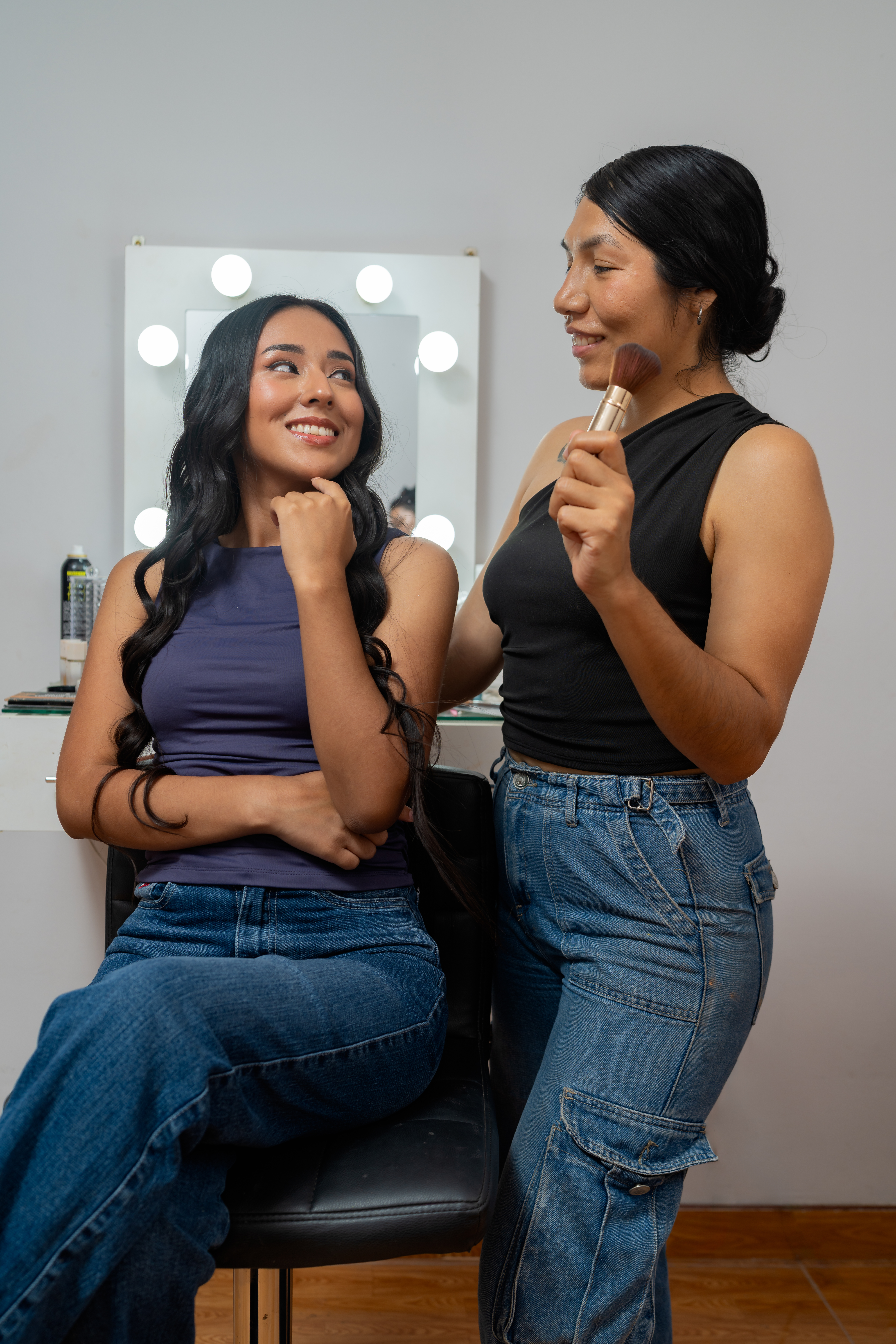 Makeup artist styling client's long dark curly hair in a beauty salon vanity mirror, both smiling during a makeover session